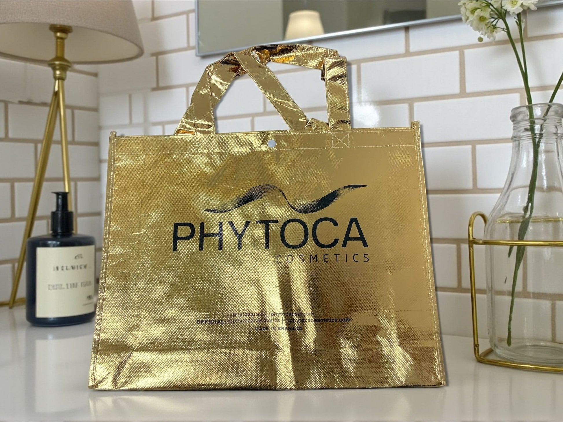 Bolsa de mano dorada metalizada Phytoca USA con logo negro impreso, ideal para cosméticos y accesorios.
