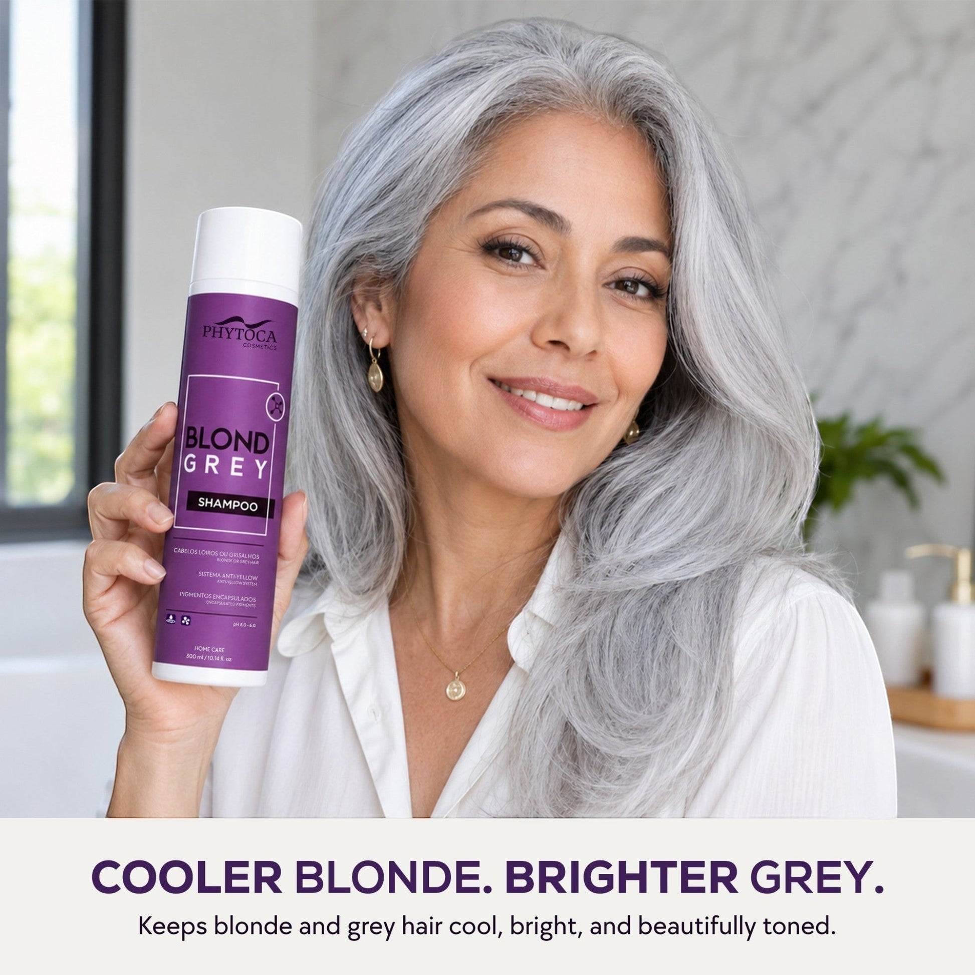 Mujer sonriente con cabello gris natural sosteniendo el champú Phytoca USA Blonde Grey para neutralizar tonos amarillos.