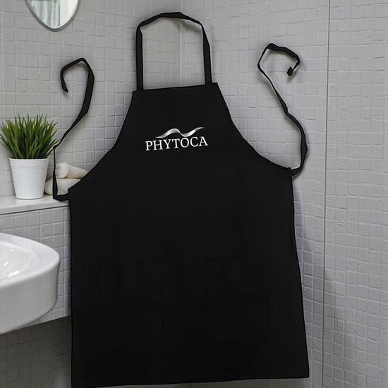 Delantal de peluquería Phytoca USA negro con bolsillos frontales y logo bordado, colgado en un baño moderno.
