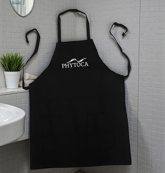 Delantal de peluquería Phytoca USA negro con bolsillos frontales y logo bordado, colgado en un baño moderno.