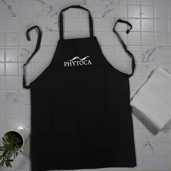 Delantal de peluquería negro Phytoca USA con bolsillos frontales y logotipo blanco bordado sobre fondo de azulejos