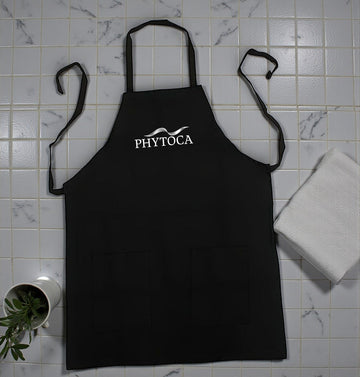 Delantal de peluquería negro Phytoca USA con bolsillos frontales y logotipo blanco bordado sobre fondo de azulejos