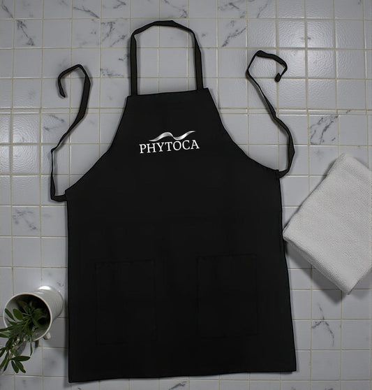 Delantal de peluquería negro Phytoca USA con bolsillos frontales y logotipo blanco bordado sobre fondo de azulejos